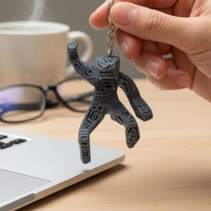 Puede incluir: Un llavero gris con forma de figura humana estilizada con un dise&ntilde;o estampado. El llavero se sostiene frente a una computadora port&aacute;til sobre un escritorio de madera. Tambi&eacute;n se ven una taza de caf&eacute; y una planta peque&ntilde;a.