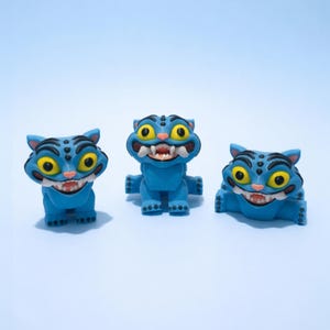 Puede incluir: Tres figuras de gato azules con ojos amarillos y bocas abiertas, cada una en una pose diferente. Las figuras tienen rayas negras y est&aacute;n sobre un fondo azul claro. Estas caprichosas figuras de gato son perfectas para coleccionistas.