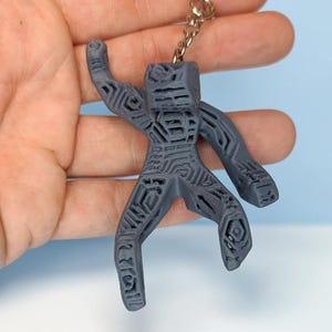Puede incluir: Un llavero gris, impreso en 3D, con forma de figura humana estilizada. La figura tiene patrones geom&eacute;tricos y est&aacute; unida a un llavero de metal. El fondo es azul claro y blanco.