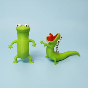 Könnte beinhalten: Zwei grüne Eidechsenfiguren mit weißen Augen und offenem Mund. Eine Eidechse steht aufrecht mit ausgestreckten Armen, während die andere eine rote Zunge und einen Schlüsselanhänger hat. Der Hintergrund ist hellblau.