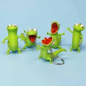 May include: Five green lizard keyrings with open red mouths and white eyes are arranged on a light blue background. One keyring has a silver ring. The lizards have their arms outstretched.