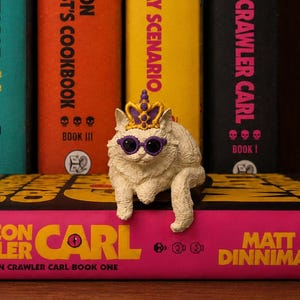 Op de afbeelding: Een wit kattenbeeldje met een paarse en gouden kroon en een paarse zonnebril, zittend op een roze boek getiteld Dungeon Crawler Carl van Matt Dinniman, met vier extra boeken van dezelfde auteur rechtopstaand op de achtergrond.