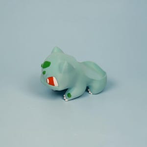 May include: A light teal Bulbasaur figurine with green and red accents. The figurine is lying down on a light blue surface. The eyes and the bulb on its back are green, and the mouth is red and white.