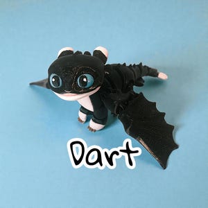 May include: A black and white dragon figurine with large blue eyes and the name "Dart" in white script. The dragon has large wings and is posed on a light blue background. The figurine is a stylized representation of a dragon.