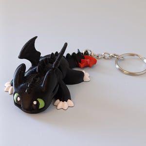 May include: A black dragon keychain with green eyes, white paws, and red accents. The dragon is lying down, attached to a silver chain and keyring. The keychain is a 3D-printed item.