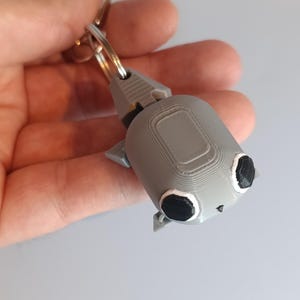 May include: A gray 3D-printed keychain featuring a stylized robot design. The robot has a rectangular body, small triangular ears, and large black eyes with white outlines. A silver key ring is attached to the top.