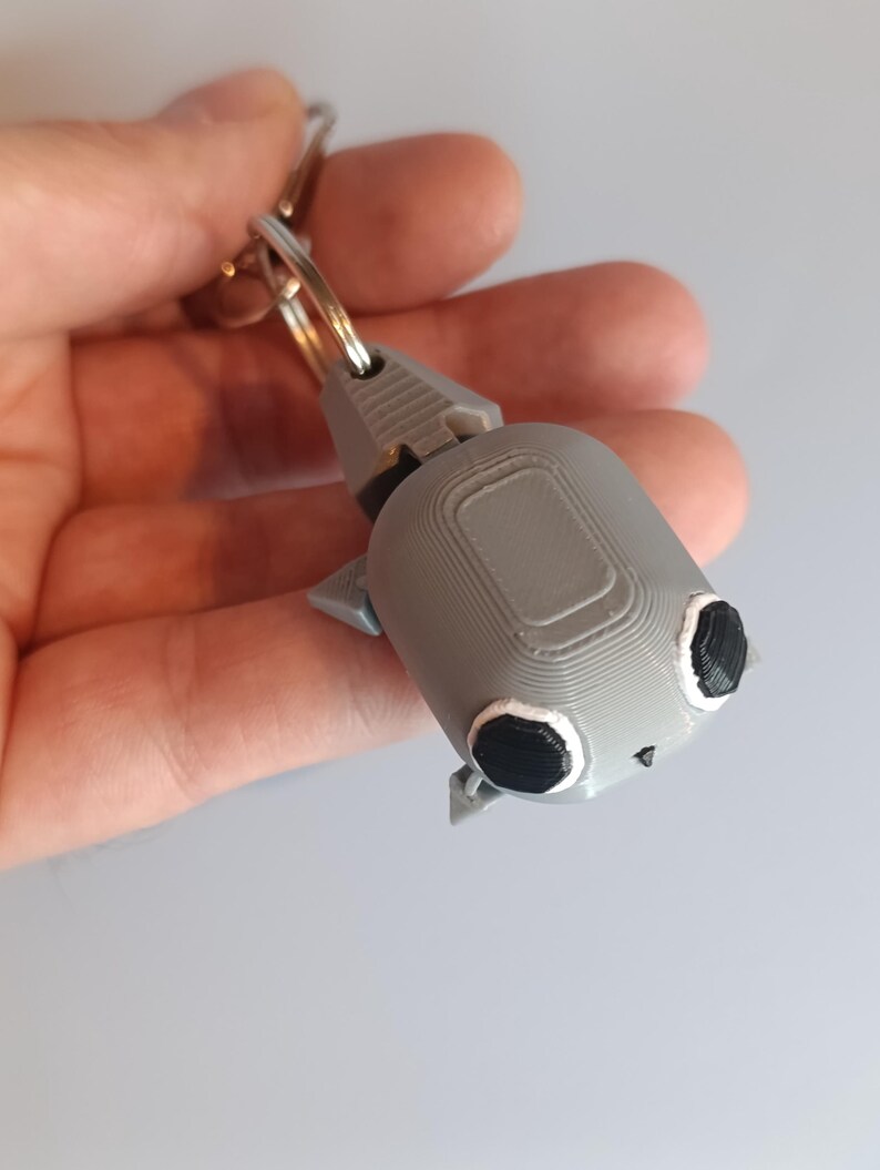 May include: A gray 3D-printed keychain featuring a stylized robot design. The robot has a rectangular body, small triangular ears, and large black eyes with white outlines. A silver key ring is attached to the top.