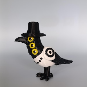 Puede incluir: Una figura de cuervo impresa en 3D en blanco y negro, con un sombrero de copa negro. El cuervo tiene tres ojos amarillos y un pecho blanco con puntos negros. El p&aacute;jaro est&aacute; de pie sobre dos patas negras.