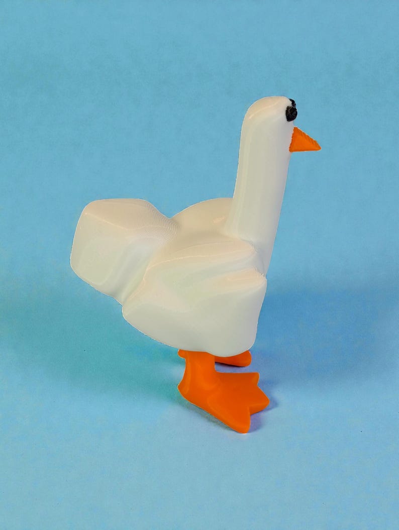 May include: A white goose figurine with orange feet and beak, and black eyes. The goose is standing on a light blue surface. The figurine has a smooth, glossy finish and is a whimsical decorative item.