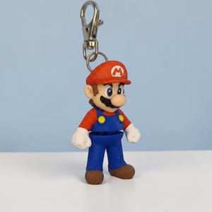 Puede incluir: Un llavero con una peque&ntilde;a y colorida figura de Mario. La figura lleva un gorro rojo con una "M" blanca, una camisa roja, un peto azul y zapatos marrones. El llavero tiene un cierre plateado. La figura mide unos 6 cm de altura.