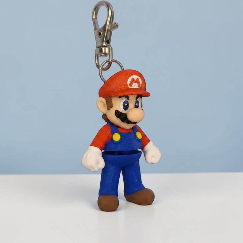Puede incluir: Un llavero con una peque&ntilde;a y colorida figura de Mario. La figura lleva un gorro rojo con una "M" blanca, una camisa roja, un peto azul y zapatos marrones. El llavero tiene un cierre plateado. La figura mide unos 6 cm de altura.