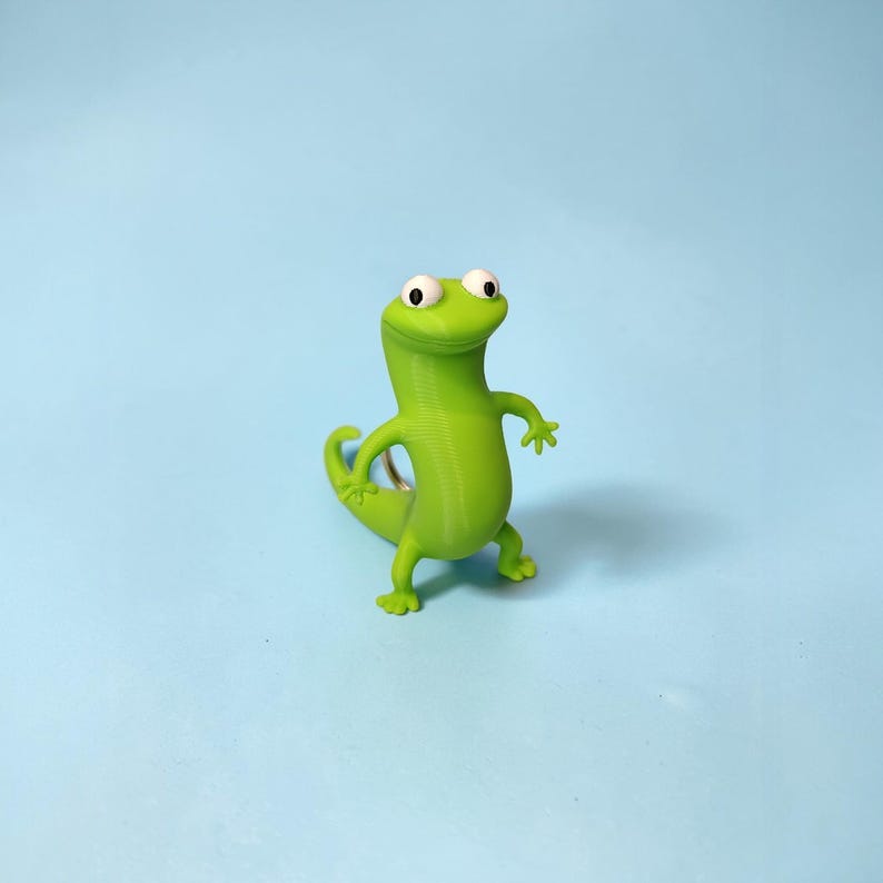 May include: A bright green, 3D-printed lizard figurine with large, white-eyed eyes and small arms and legs. The lizard is posed as if it is walking or dancing. The background is a solid light blue.