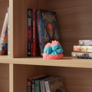Puede incluir: Una peque&ntilde;a figura impresa en 3D de un drag&oacute;n durmiente con cuerpo azul claro y detalles en rosa, descansando dentro de un nido rosa texturizado. El drag&oacute;n tiene los ojos cerrados y un acabado mate suave, colocado en un estante de madera entre libros.