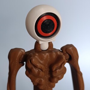 May include: A 3D-printed figurine featuring a large, white, spherical head with a black and red eye design. The body is brown with textured details, and the arms are outstretched. The figure has a unique, stylized appearance.