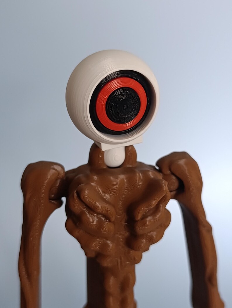 May include: A 3D-printed figurine featuring a large, white, spherical head with a black and red eye design. The body is brown with textured details, and the arms are outstretched. The figure has a unique, stylized appearance.