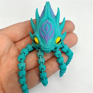 May include: A teal articulated creature toy with a purple and teal head and yellow eyes. The toy has a segmented body and tentacles. The toy is held in a hand.