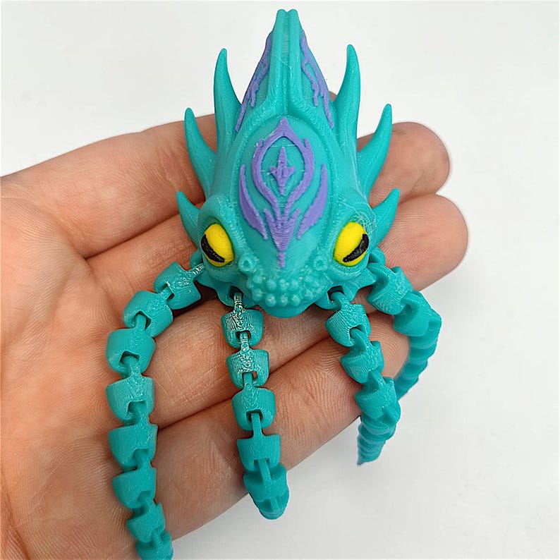 May include: A teal articulated creature toy with a purple and teal head and yellow eyes. The toy has a segmented body and tentacles. The toy is held in a hand.