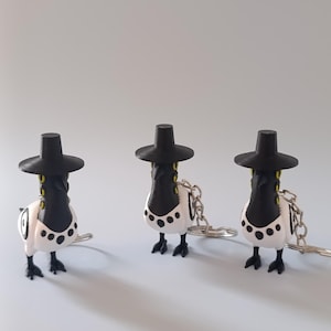 May include: Three novelty keychains featuring a bird-like design. Each keychain has a black hat, a black and white body with black spots, and a silver-colored chain. The keychains are standing on a light gray surface.