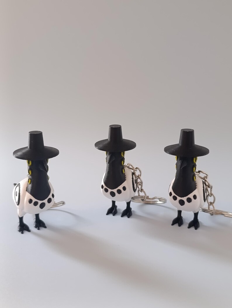 May include: Three novelty keychains featuring a bird-like design. Each keychain has a black hat, a black and white body with black spots, and a silver-colored chain. The keychains are standing on a light gray surface.