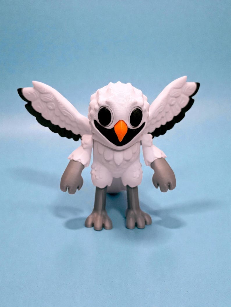 May include: A white owl figurine with outstretched wings, black accents, and an orange beak. The owl has large, round eyes and gray feet and hands. The figurine is set against a light blue background.