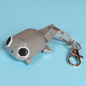 May include: A gray, 3D-printed keychain shaped like a stylized tadpole. The keychain features a silver clasp and ring for attaching to keys or bags. The tadpole has black eyes and small fins. The background is a solid light blue.