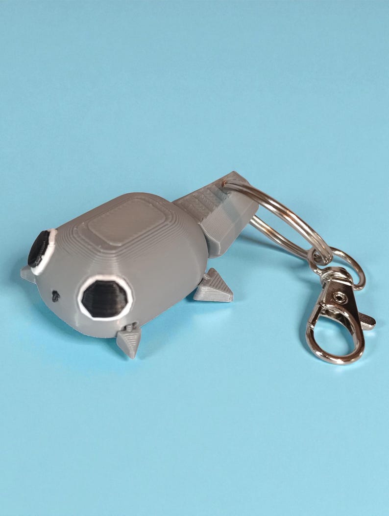 May include: A gray, 3D-printed keychain shaped like a stylized tadpole. The keychain features a silver clasp and ring for attaching to keys or bags. The tadpole has black eyes and small fins. The background is a solid light blue.