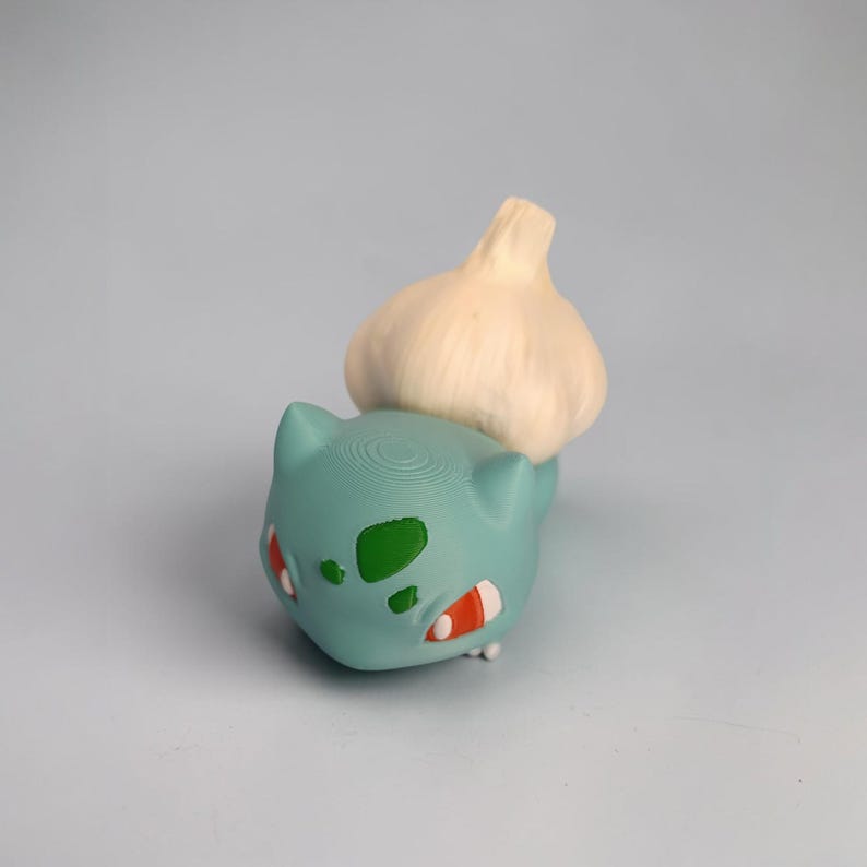 May include: A teal Bulbasaur figure with a white garlic bulb on its back. The Bulbasaur has green spots, red and white accents, and a textured surface. The figure is a unique, whimsical design.