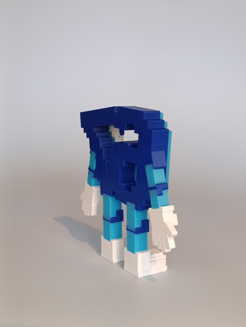 Pode incluir: Uma figura impressa em 3D com um design em blocos. A figura &eacute; principalmente azul, com pernas azul claro e m&atilde;os e p&eacute;s brancos. A figura tem uma est&eacute;tica geom&eacute;trica e pixelizada, sugerindo um tema digital ou retro.