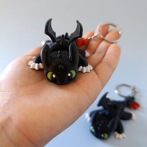 May include: Black dragon keychain with green eyes and white accents. The dragon has a red tail accent and is attached to a silver keyring. A second dragon keychain is visible in the background.