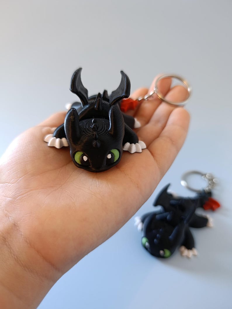 May include: Black dragon keychain with green eyes and white accents. The dragon has a red tail accent and is attached to a silver keyring. A second dragon keychain is visible in the background.