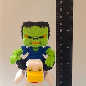 May include: A 3D-printed figurine of a green-skinned character with black hair and a blue shirt riding a white and yellow duck. The figurine is approximately 4 inches tall, with a ruler in the background for scale.