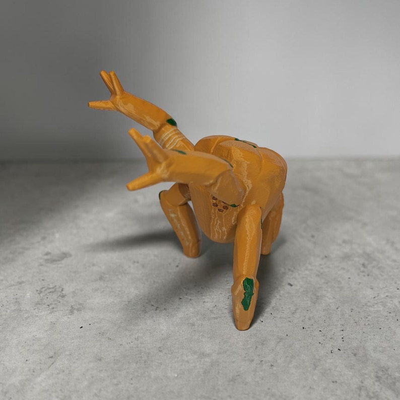 May include: A 3D-printed orange insect-like figurine with green accents on its legs and back. The figurine is posed in a crawling position, with its arms raised. The object is on a gray surface.
