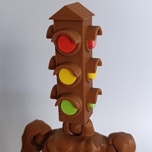 May include: A brown 3D-printed sculpture of a traffic light with a humanoid body. The traffic light head has red, yellow, and green lights. The figure has arms and a textured surface.