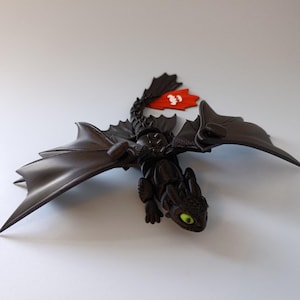 K&ouml;nnte beinhalten: Eine schwarze Drachenfigur mit gro&szlig;en Fl&uuml;geln und einer roten Flosse. Der Drache hat gr&uuml;ne Augen und einen detaillierten, strukturierten K&ouml;rper. Eine rote Flosse mit wei&szlig;en Markierungen ist am R&uuml;cken des Drachen befestigt.