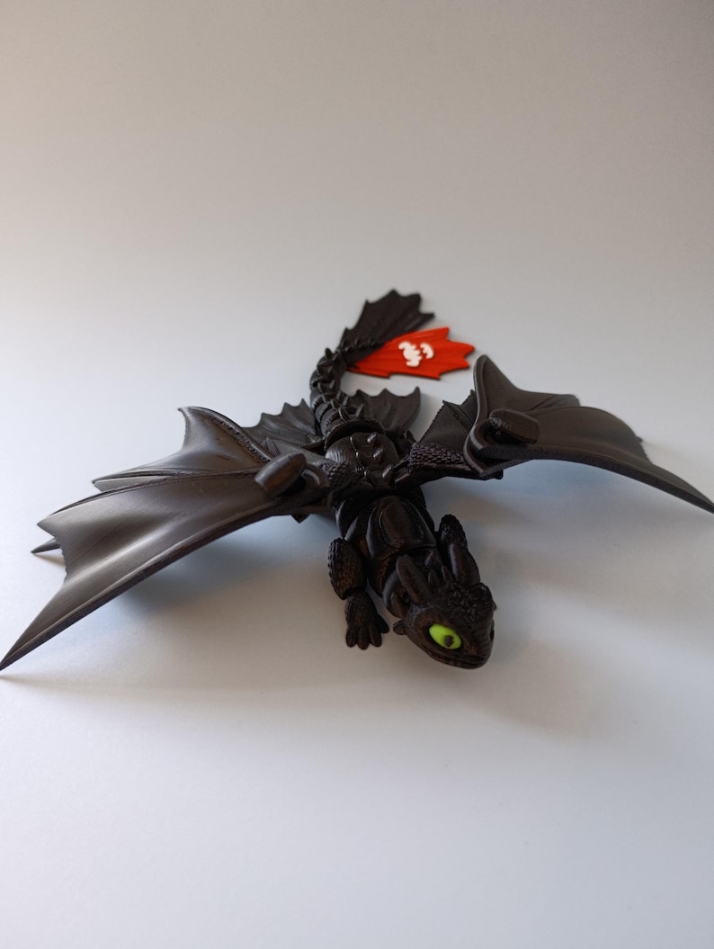 K&ouml;nnte beinhalten: Eine schwarze Drachenfigur mit gro&szlig;en Fl&uuml;geln und einer roten Flosse. Der Drache hat gr&uuml;ne Augen und einen detaillierten, strukturierten K&ouml;rper. Eine rote Flosse mit wei&szlig;en Markierungen ist am R&uuml;cken des Drachen befestigt.