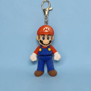 Puede incluir: Un llavero colorido con una figura en miniatura de Mario, un popular personaje de videojuegos. La figura lleva una gorra roja con el logo 'M' blanco, una camisa roja, un peto azul y zapatos marrones. El llavero tiene un cierre plateado.