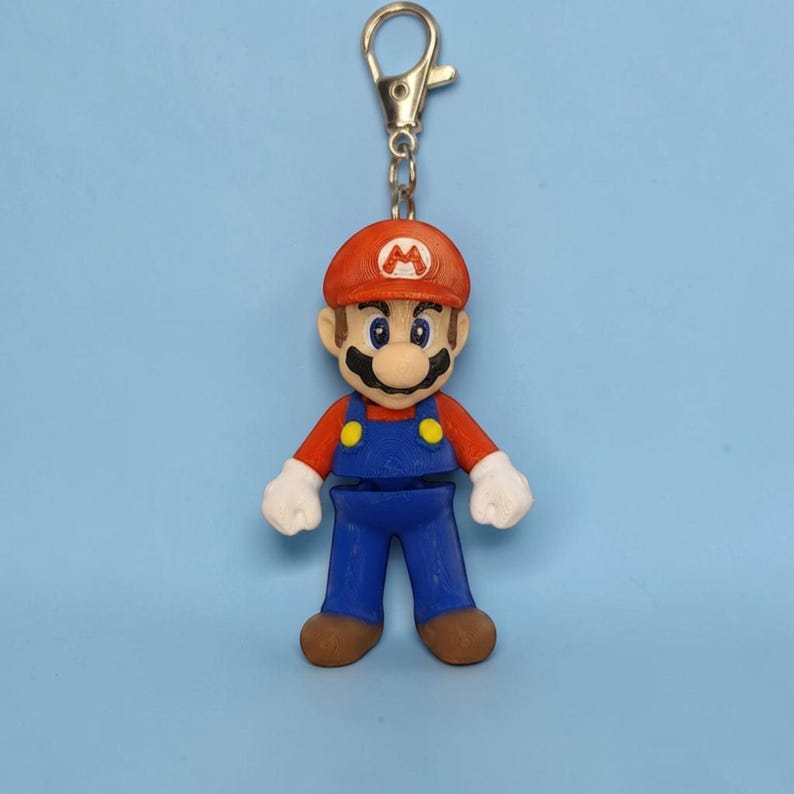 Puede incluir: Un llavero colorido con una figura en miniatura de Mario, un popular personaje de videojuegos. La figura lleva una gorra roja con el logo 'M' blanco, una camisa roja, un peto azul y zapatos marrones. El llavero tiene un cierre plateado.