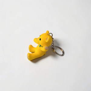 Puede incluir: Un llavero de oso de color amarillo brillante con un acabado mate suave, un peque&ntilde;o accesorio de cuello en color verde azulado y una anilla de metal plateado unida a la parte superior. El oso aparece sentado con orejas redondas y ojos oscuros.