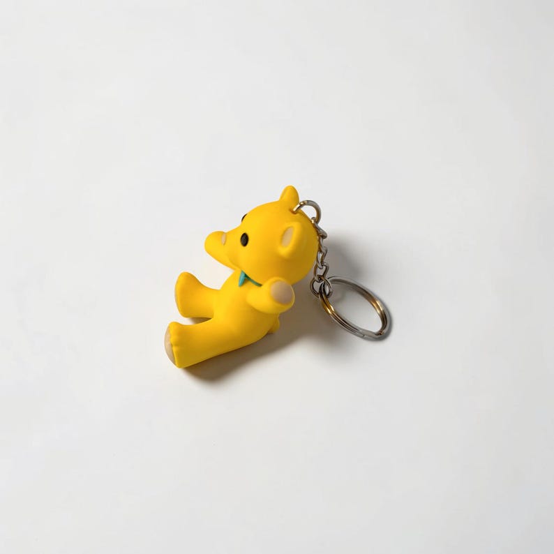 Puede incluir: Un llavero de oso de color amarillo brillante con un acabado mate suave, un peque&ntilde;o accesorio de cuello en color verde azulado y una anilla de metal plateado unida a la parte superior. El oso aparece sentado con orejas redondas y ojos oscuros.