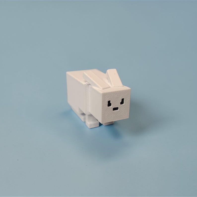 Op de afbeelding: Een witte, blokkerige konijnenfiguur met een minimalistisch ontwerp. Het konijn heeft een rechthoekig lichaam, een vierkante kop en kleine, rechtopstaande oren. De gezichtskenmerken zijn eenvoudige zwarte oog- en monddetails. De figuur staat tegen een lichtblauwe achtergrond.