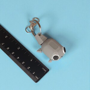 May include: A gray robot-shaped keychain with black eye details and a silver clasp. The keychain is next to a black ruler with inch and millimeter measurements. The background is a solid light blue.