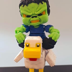 May include: A 3D-printed toy featuring a green-skinned monster riding a white and yellow pixelated chicken. The monster has black hair, a blue shirt, and an angry expression. The chicken has a red wattle.