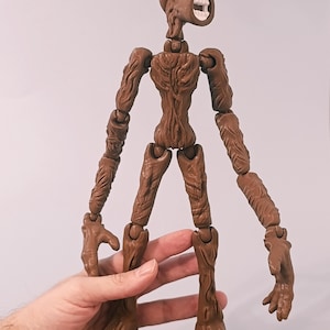 Op de afbeelding: Een bruin Siren Head actiefiguur met beweegbare gewrichten. De figuur heeft een getextureerd, boomachtig uiterlijk, met een hoofd in de vorm van twee luidsprekers. Het figuur wordt door een hand tegen een effen achtergrond gehouden.