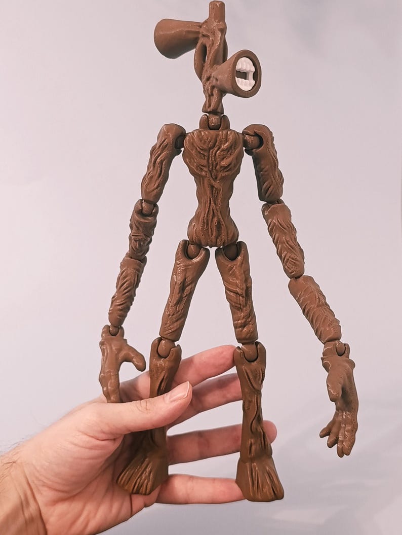 Op de afbeelding: Een bruin Siren Head actiefiguur met beweegbare gewrichten. De figuur heeft een getextureerd, boomachtig uiterlijk, met een hoofd in de vorm van twee luidsprekers. Het figuur wordt door een hand tegen een effen achtergrond gehouden.