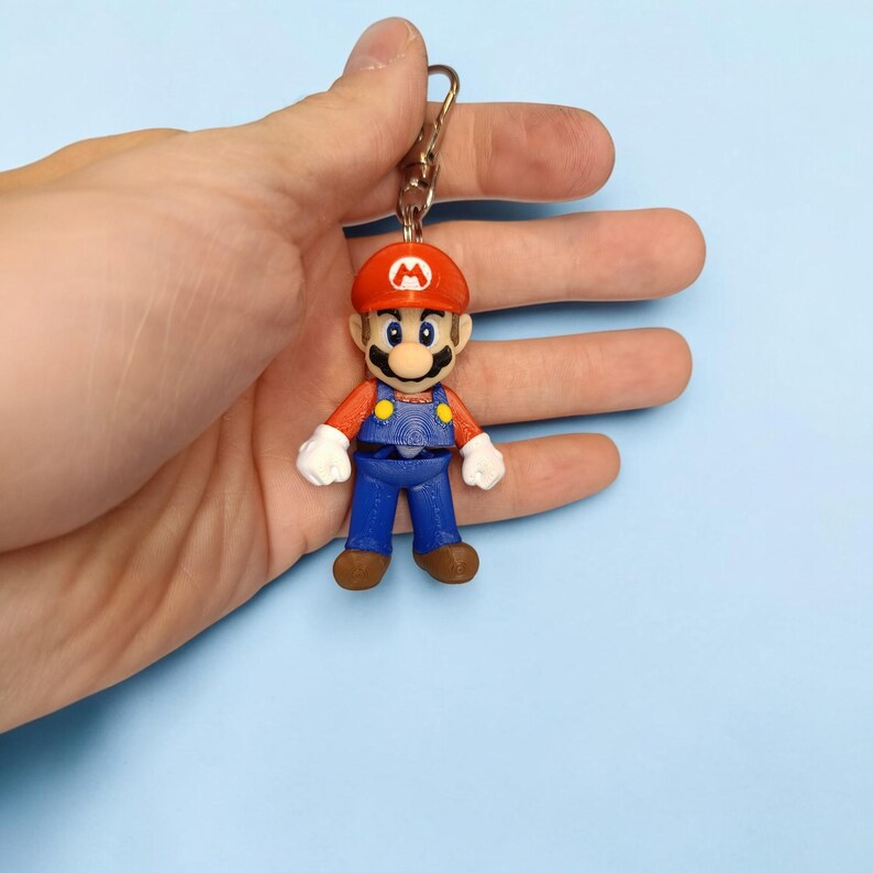 Puede incluir: Un llavero impreso en 3D con una figura en miniatura de Mario. La figura viste su gorra roja caracter&iacute;stica, un peto azul y una camisa roja. El llavero tiene un cierre met&aacute;lico dorado.