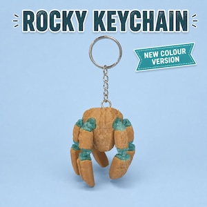 May include: A Rocky keychain featuring a tan and teal crab-shaped charm attached to a silver metal split ring and chain. The text ROCKY KEYCHAIN appears at the top, and a teal banner with the text NEW COLOUR VERSION is positioned to the right.
