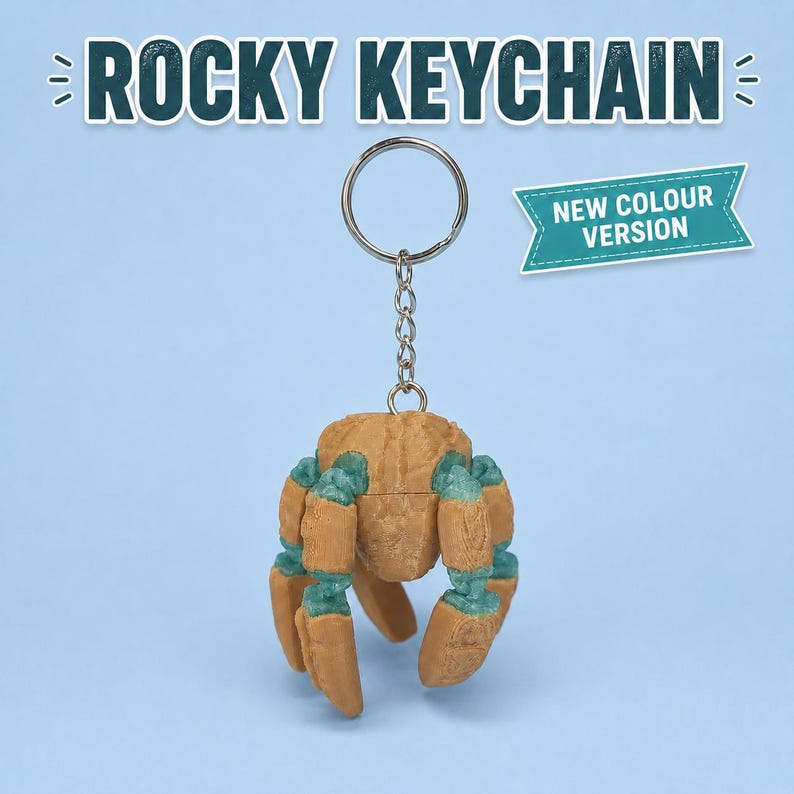 May include: A Rocky keychain featuring a tan and teal crab-shaped charm attached to a silver metal split ring and chain. The text ROCKY KEYCHAIN appears at the top, and a teal banner with the text NEW COLOUR VERSION is positioned to the right.