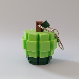 Puede incluir: Un llavero verde en forma de manzana pixelada. El llavero est&aacute; hecho de peque&ntilde;os bloques cuadrados en tonos de verde y verde oscuro. Tiene un tallo marr&oacute;n y un llavero plateado.