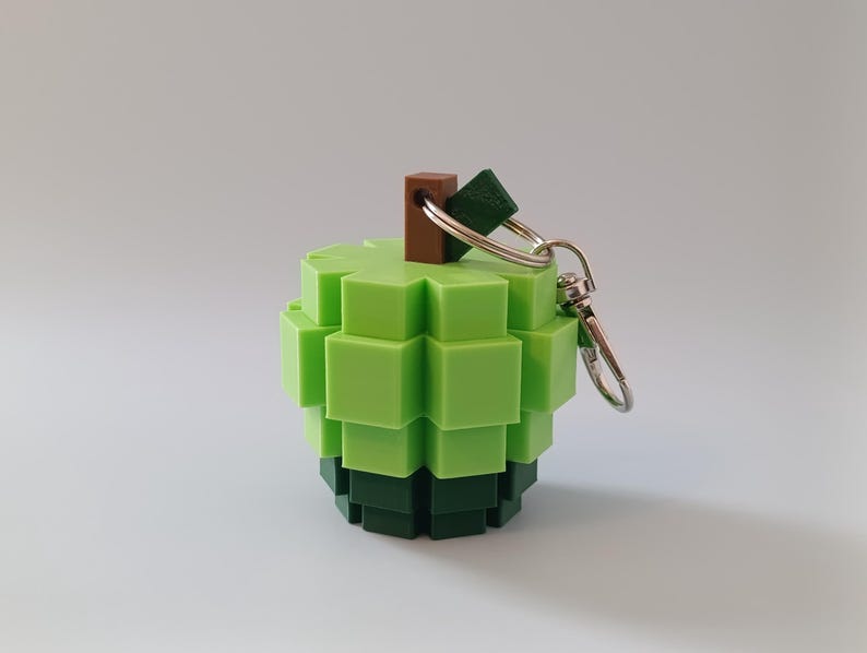 Puede incluir: Un llavero verde en forma de manzana pixelada. El llavero est&aacute; hecho de peque&ntilde;os bloques cuadrados en tonos de verde y verde oscuro. Tiene un tallo marr&oacute;n y un llavero plateado.