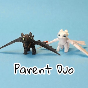 May include: Two dragon figurines, one black and one white, with outstretched wings, stand on a blue background. The black dragon has green eyes and the white dragon has blue eyes. The text "Parent Duo" is at the bottom.